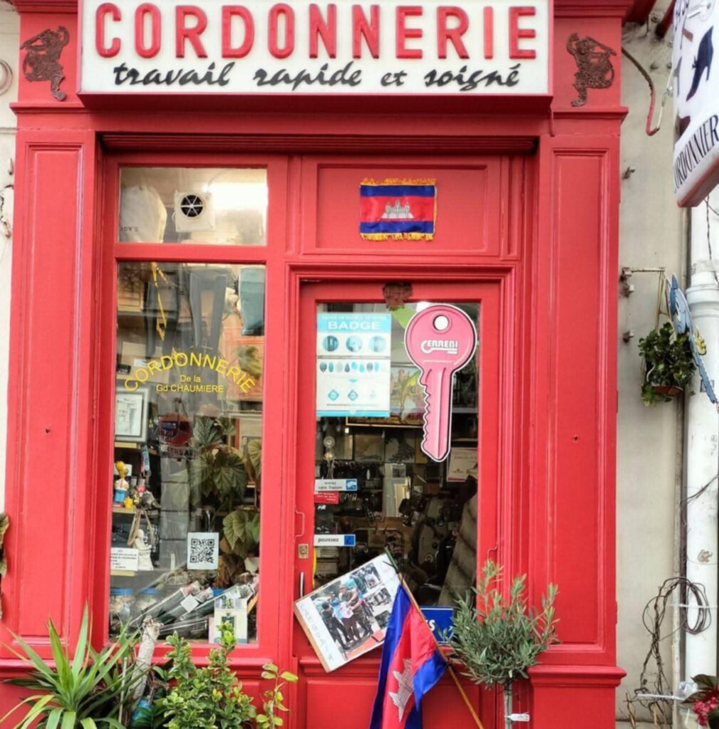 Façade rouge de la Cordonnerie de la Grande Chaumière à Paris, avec un drapeau cambodgien affiché et des outils d’artisan cordonnier visibles dans la vitrine.