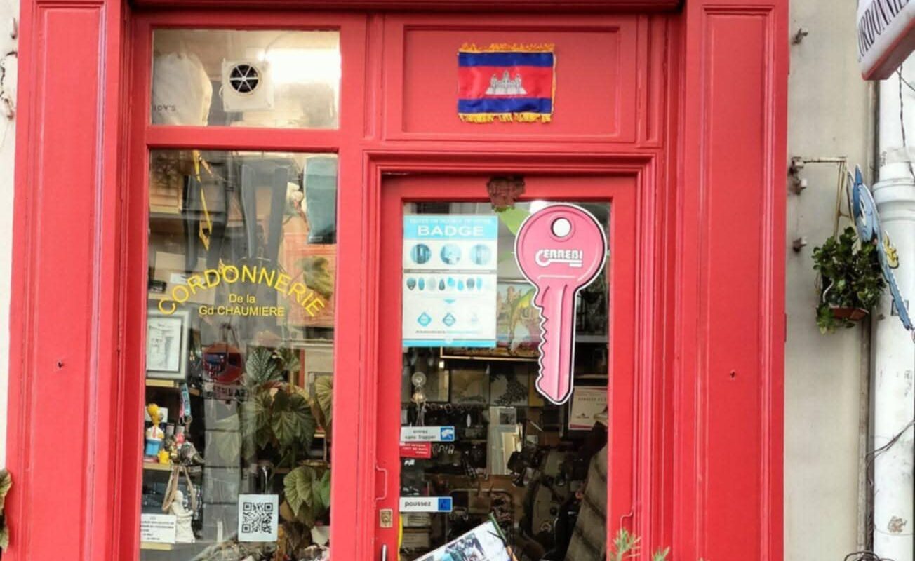 Façade rouge de la Cordonnerie de la Grande Chaumière à Paris, avec un drapeau cambodgien affiché et des outils d’artisan cordonnier visibles dans la vitrine.