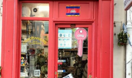 Façade rouge de la Cordonnerie de la Grande Chaumière à Paris, avec un drapeau cambodgien affiché et des outils d’artisan cordonnier visibles dans la vitrine.