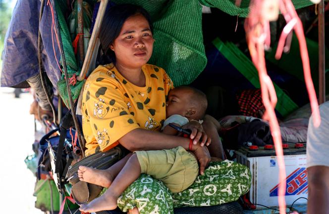 Une femme assise dans un abri de fortune tient un enfant endormi contre elle. Des bâches, sacs et effets personnels l’entourent, témoignant d’un déplacement et de conditions de vie précaires.