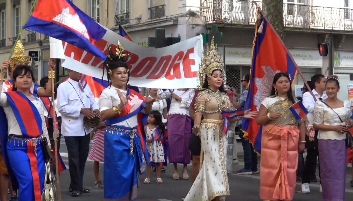 Défilé de la communauté cambodgienne à Lyon lors de la Fête des Bannières du Monde. Des femmes et des enfants portent des costumes traditionnels khmers, des couronnes ornées et des drapeaux du Cambodge, avançant ensemble dans la rue, symbole de fierté culturelle et de transmission.