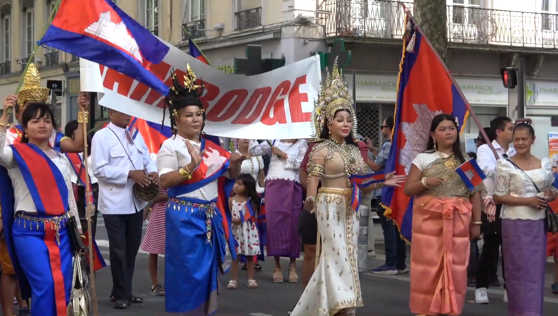 Défilé de la communauté cambodgienne à Lyon lors de la Fête des Bannières du Monde. Des femmes et des enfants portent des costumes traditionnels khmers, des couronnes ornées et des drapeaux du Cambodge, avançant ensemble dans la rue, symbole de fierté culturelle et de transmission.